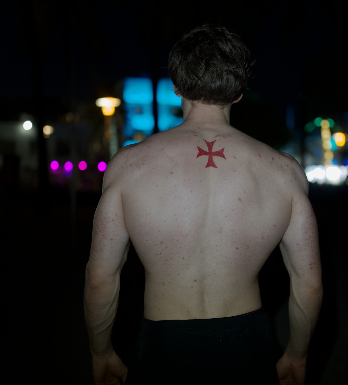 Collin Christiansen photographed from behind at night, red cross tattoo on his upper back, city lights blurred behind him